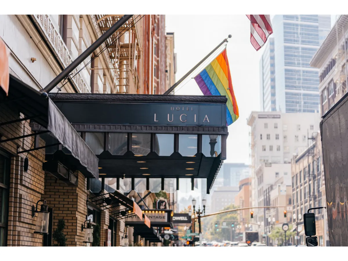 The exterior of the hotel is adorned with a rainbow flag and American flag, signaling inclusivity and community engagement. The hotel sign is visible, and the street scene hints at a lively, walkable area perfect for heading out on foot or bicycle. The atmosphere is energetic, reflecting the accessibility and vibrancy described in the section.