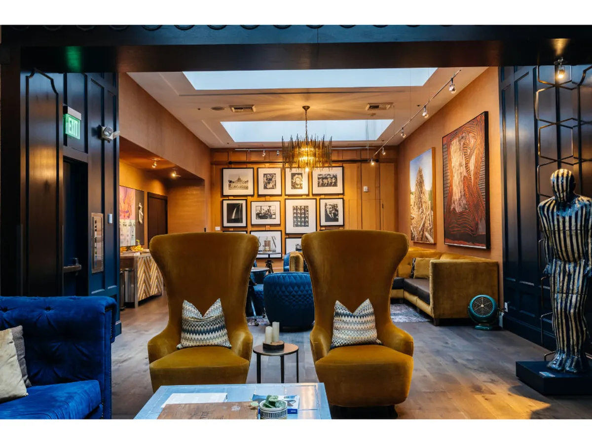 A sophisticated lounge space with yellow armchairs, a blue sofa, dramatic chandelier lighting, and curated art on the walls. The atmosphere is both artistic and welcoming, suggesting a space for art appreciation, casual drinks, and lively conversation.