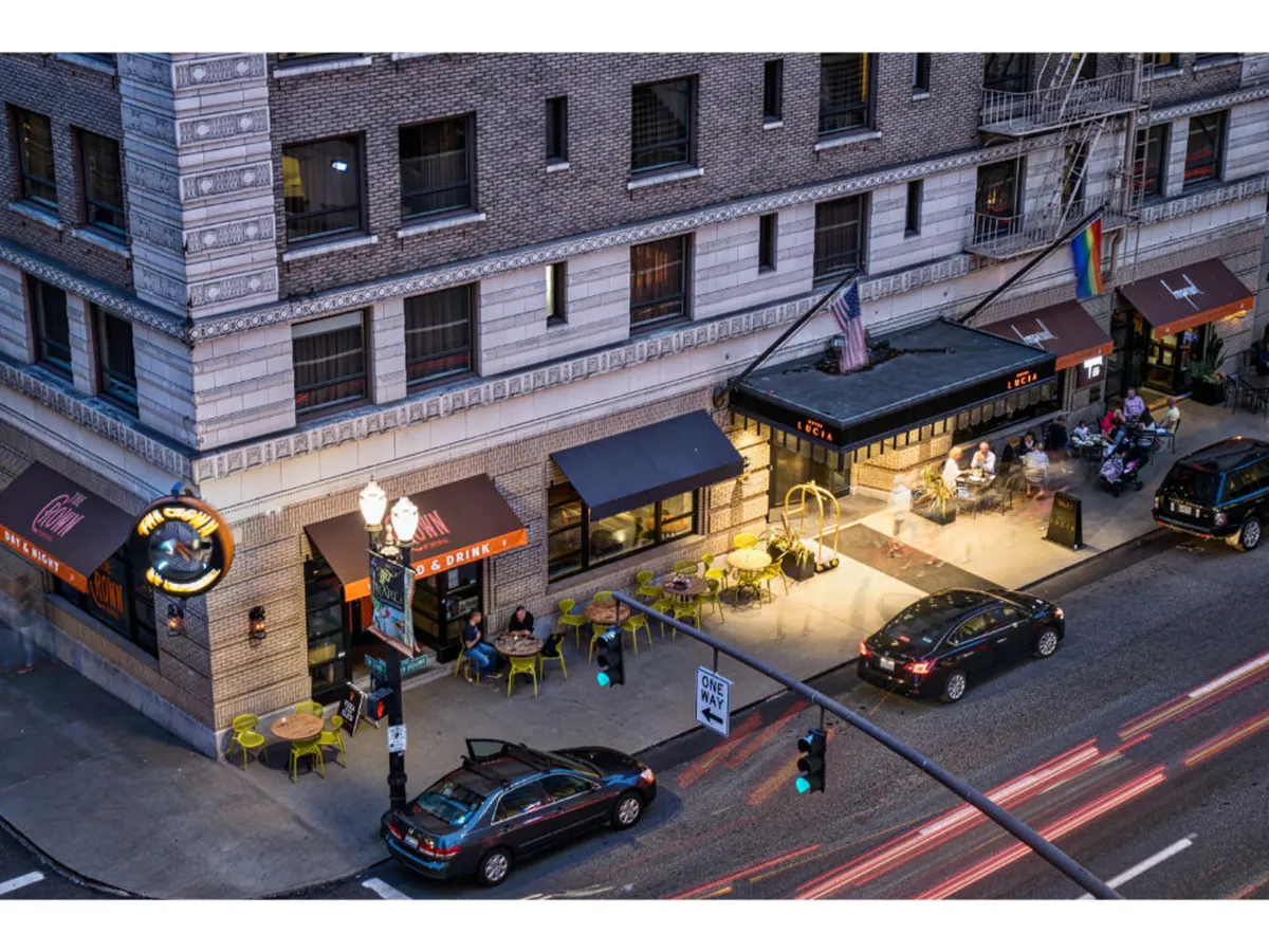 The image showcases a lively, well-lit outdoor restaurant scene with bustling street activity, illuminated signage, and a welcoming entrance. People are dining al fresco, socializing, and enjoying the city’s nightlife, embodying the social spirit of Portland.
