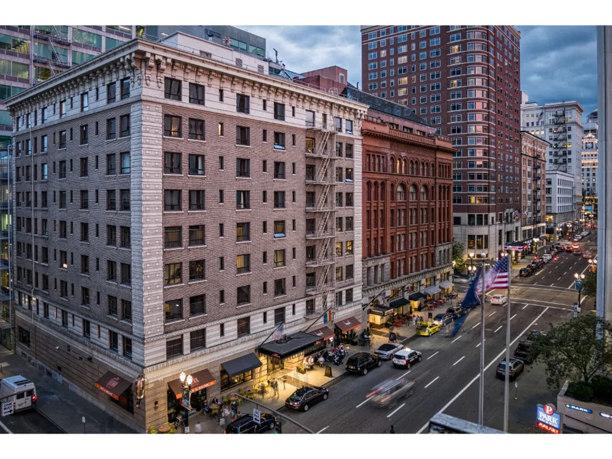 A bustling city street with historic buildings, outdoor dining areas, waving flags, and glimpses of the city skyline. People are visible walking and enjoying the urban surroundings, evoking the dynamic city life and cultural opportunities accessible from the hotel.