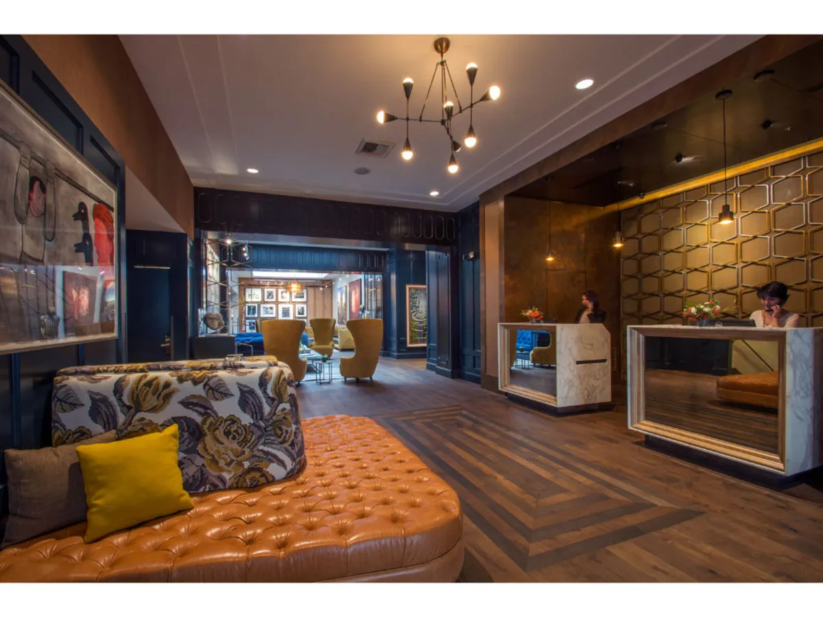A stylish, contemporary lobby area featuring a textured orange sofa, geometric reception desk, modern artwork on the walls, and unique lighting fixtures. The space buzzes with social activity while inviting guests to relax, check in, or admire the art displays.