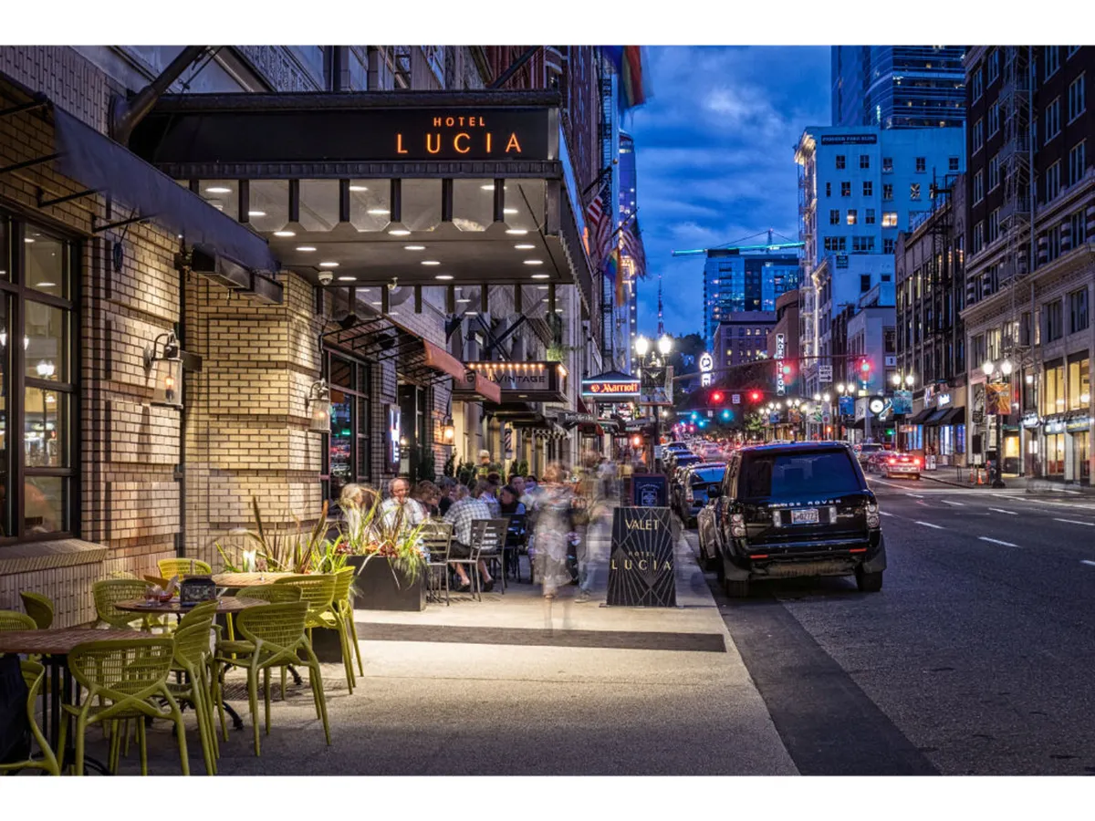 This exterior shot captures the Hotel Lucia sign, an inviting outdoor seating area, and street lights illuminating a vibrant sidewalk scene. The environment feels energetic with hints of nearby nightlife and urban pulse, directly evoking the feeling of being at the heart of downtown Portland.