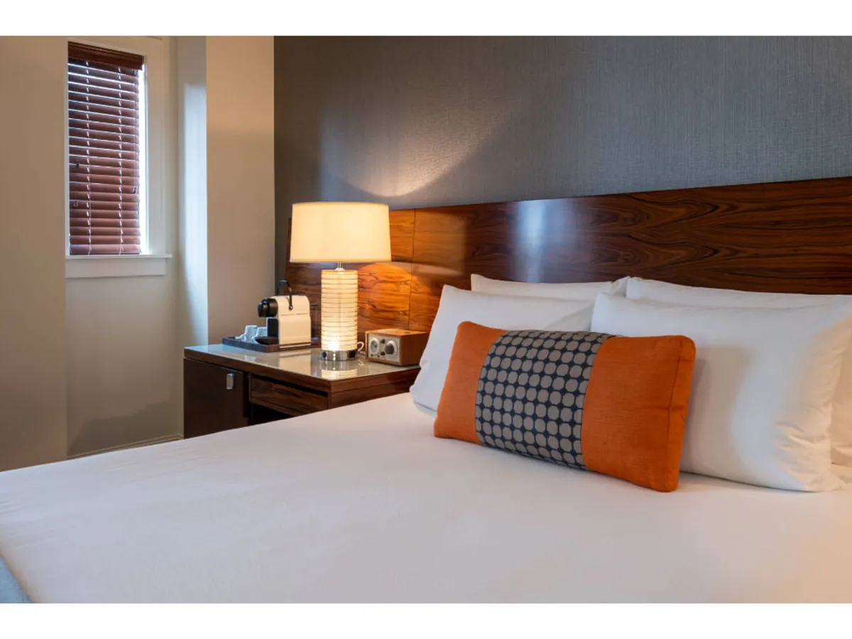 A Deluxe King room featuring a plush king-size bed with crisp white linens and a decorative pillow. Soft lighting from a bedside lamp creates a soothing, welcoming atmosphere, and a nightstand adds practicality. The overall mood is comfortable and inviting, ideal for relaxing or a restful night.