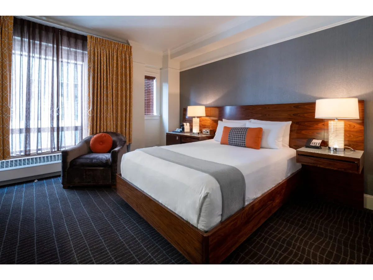 The image showcases a stylish hotel room featuring a plush bed with crisp linens, elegant nightstands, soft curtains filtering natural light, and a modern armchair inviting relaxation. The room exudes warmth and comfort, with thoughtful touches that create a cozy urban sanctuary.