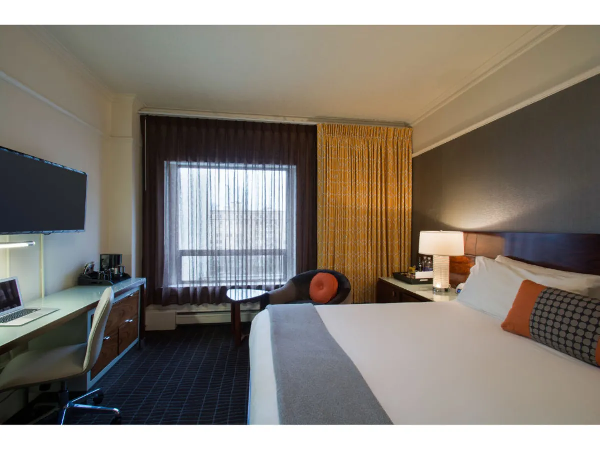 A Deluxe King room with a neatly made bed, a large window letting in natural daylight, a desk and chair for quiet work or reflection, and thick curtains for privacy. The room feels peaceful and offers views to the outside, emphasizing the retreat aspect after city adventures.